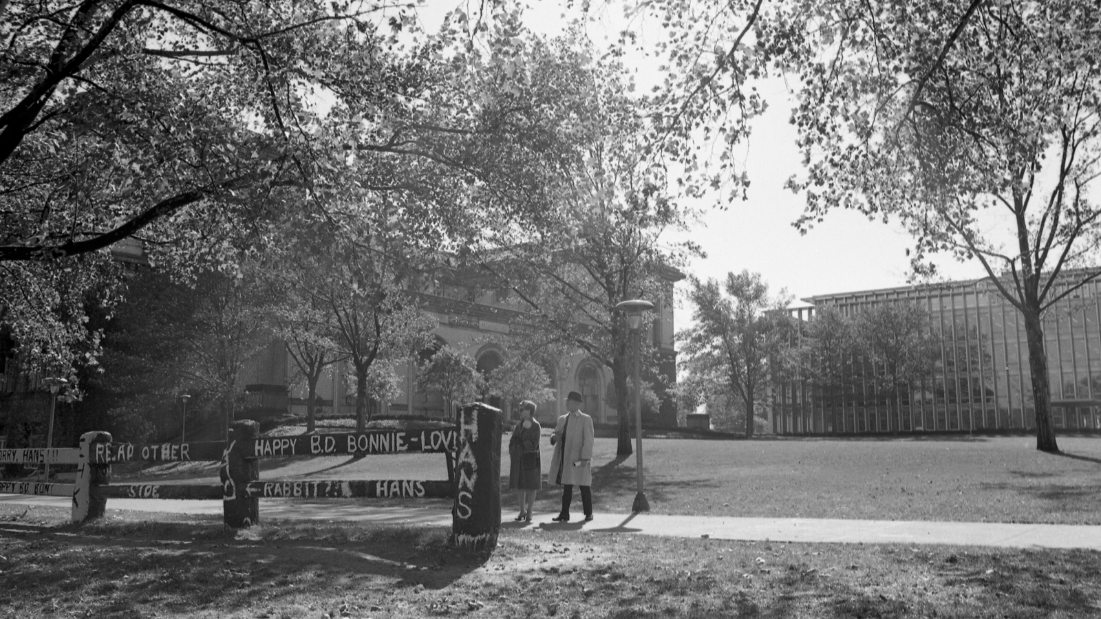 Black and white photo of two people stand outside by the decorating fence on the quad.