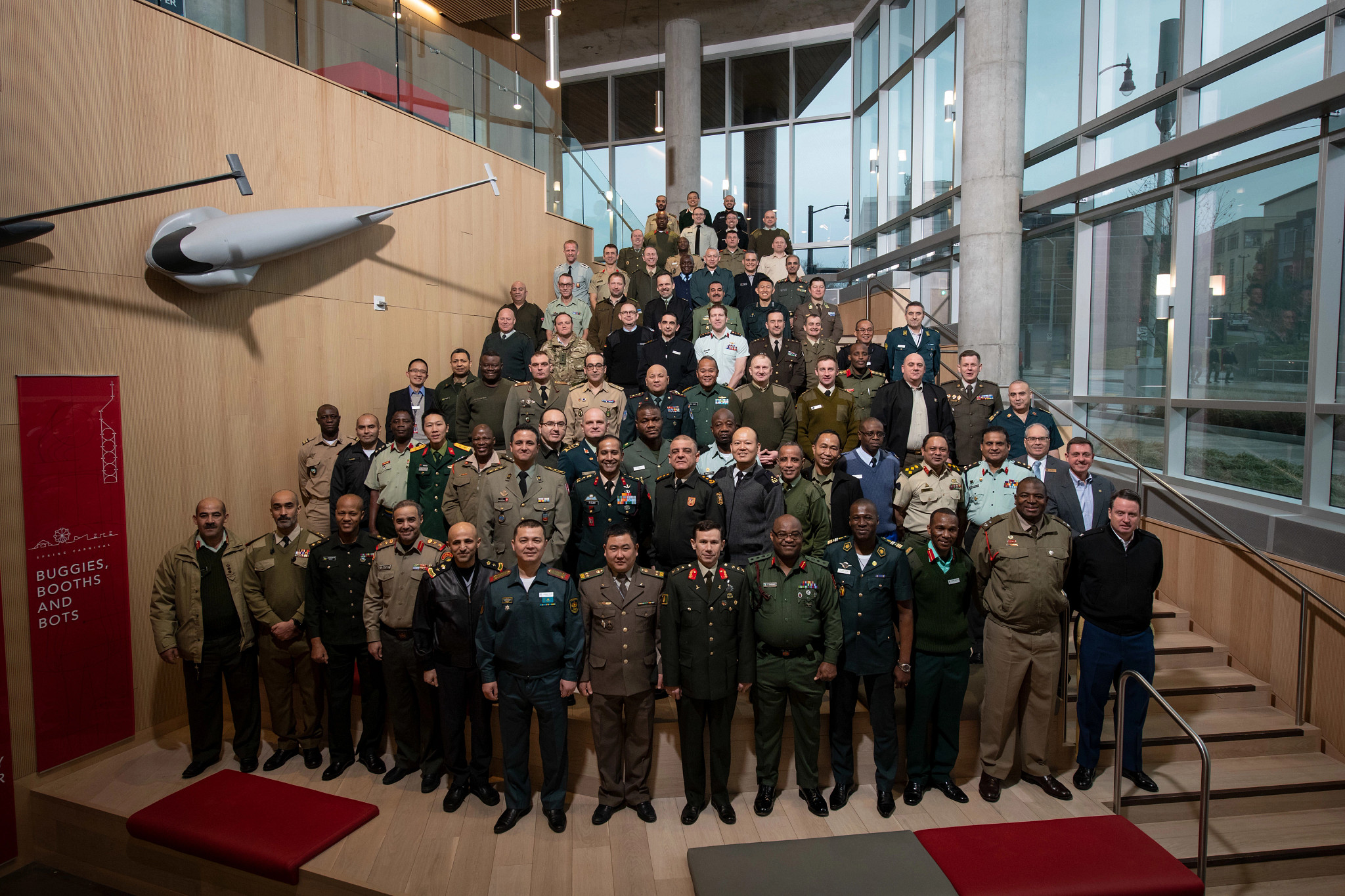 A large group of military personnel in diverse uniforms stand on a staircase in a modern building with large windows, conveying unity and professionalism.