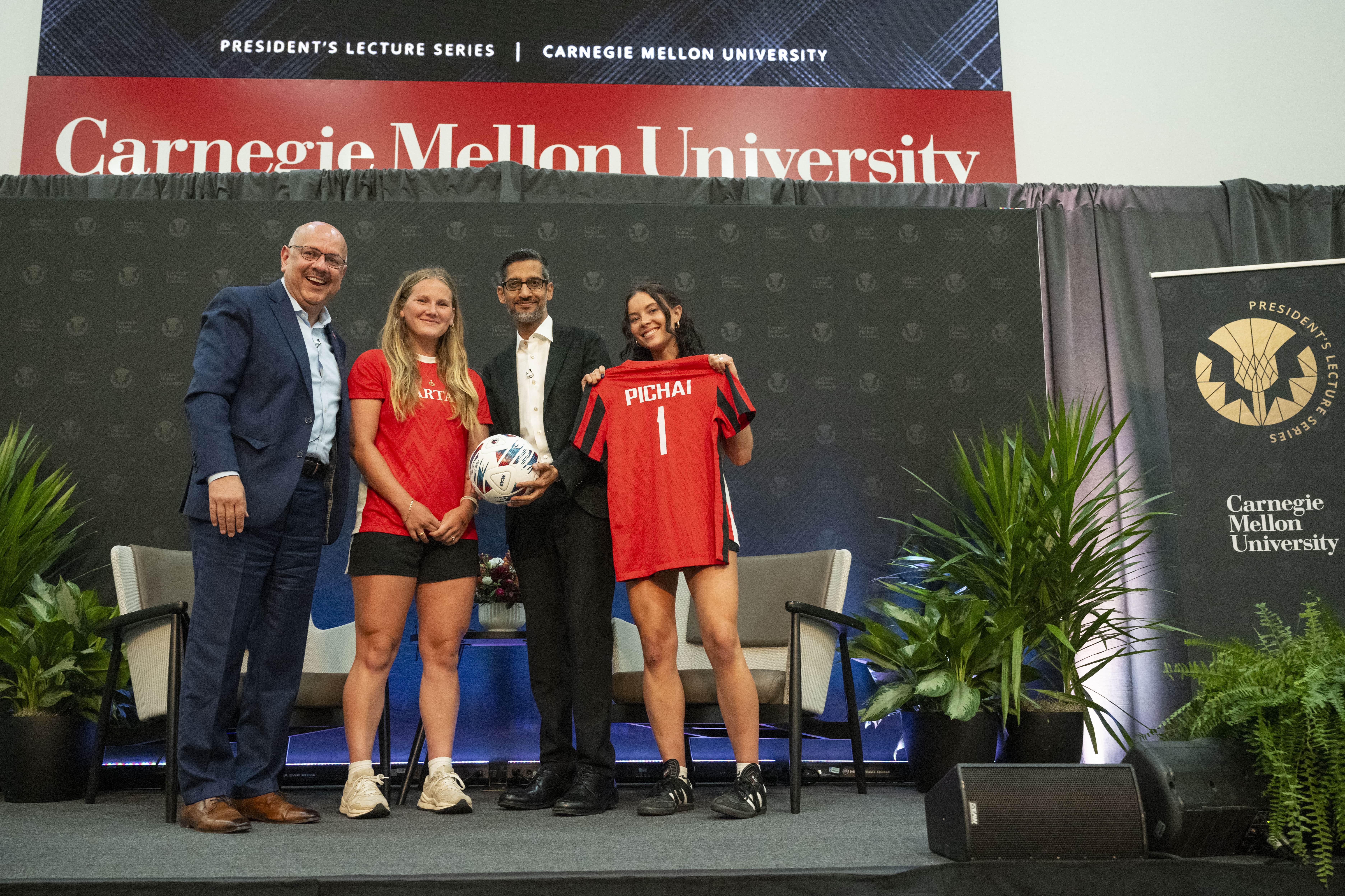 Pichai holds a soccer ball and stands next to Jahanian and two soccer players wearing red jerseys.