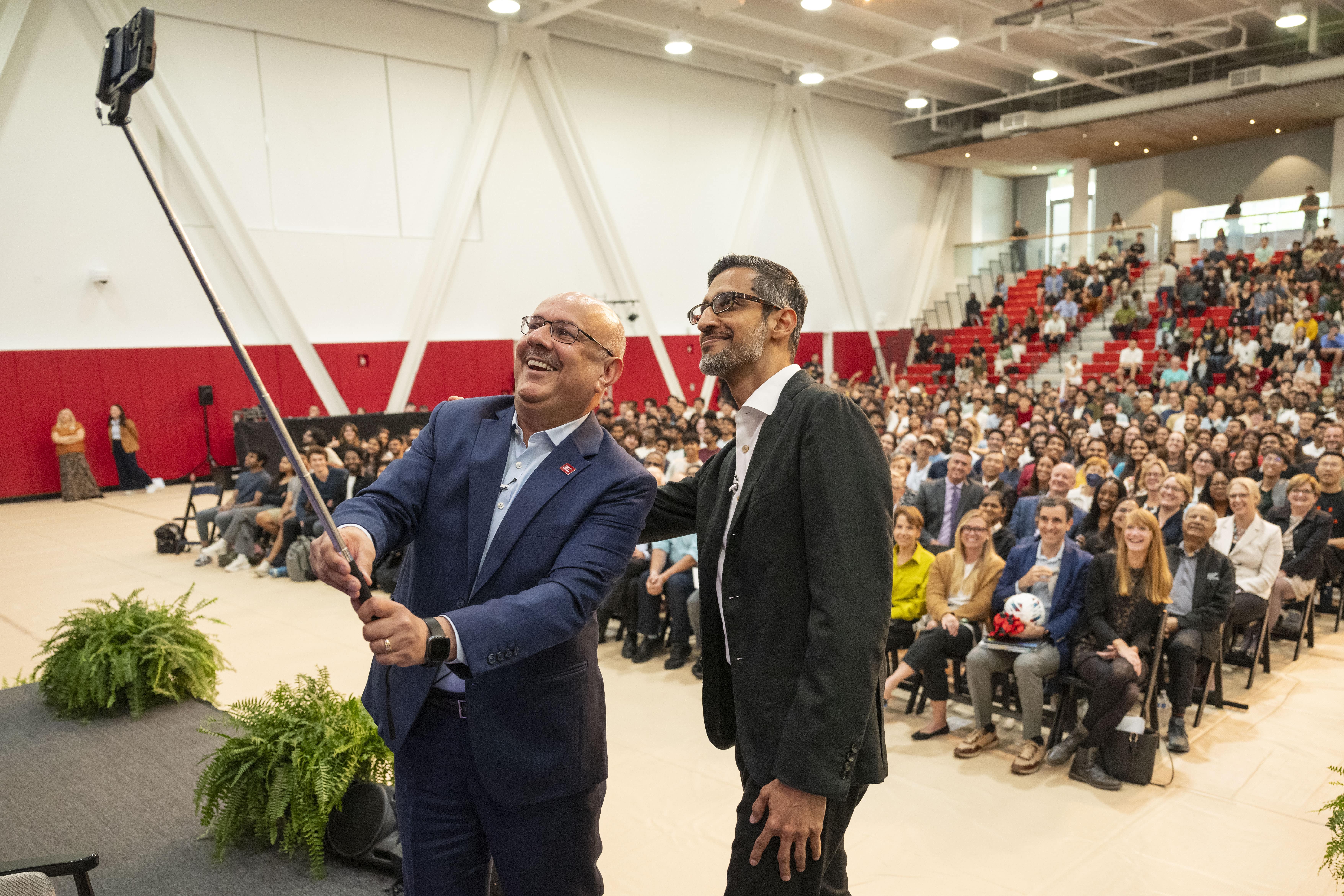 Jahanian and Pichai smile into a camera on a selfie stick. They have their back to a smiling audience.