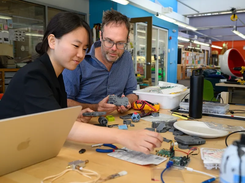 Student and professor interact with colorful robotic pieces on a wooden table.
