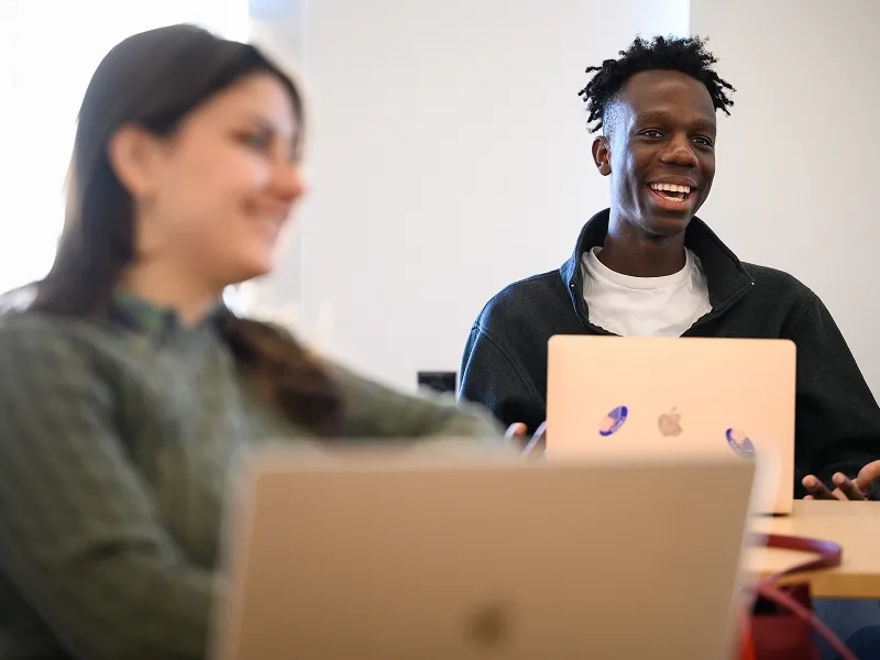 Two students smile as they type together on a silver laptop.