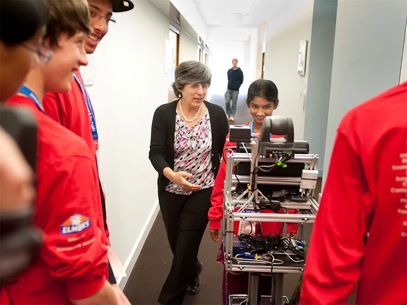 Young student pushing around tech on a cart with CMU teacher nearby.