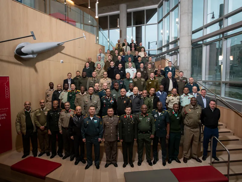 A large group of military personnel in diverse uniforms stand on a staircase in a modern building with large windows, conveying unity and professionalism.