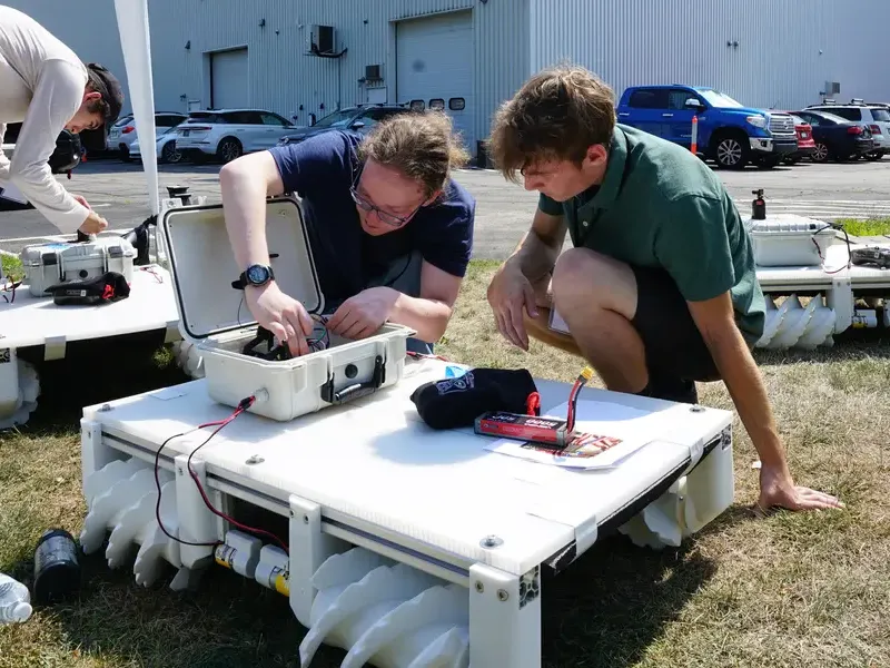 Two students assemble a robot in front of a building labeled NREC