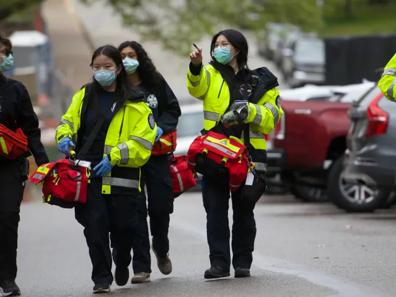 A group of focused paramedics in yellow jackets, face masks, and gloves walk down a residential street, carrying red medical bags. One gestures ahead.