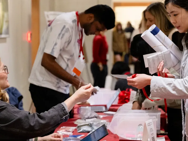 A busy registration table at a conference with attendees receiving materials. A staff member hands a badge to a woman holding rolled papers.
