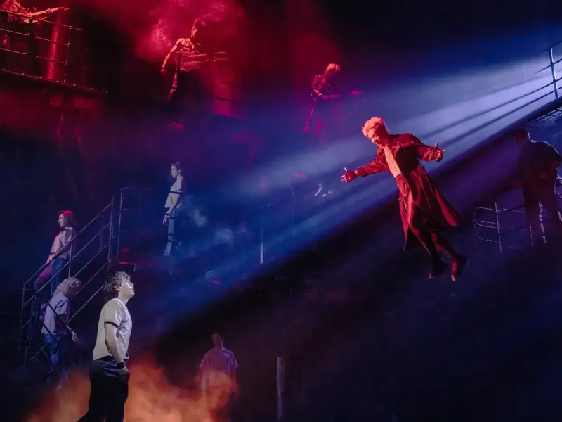 A theatrical scene with a man in a long coat suspended mid-air, bathed in blue light. Below, another person gazes up amidst a dramatic red-lit setting. Mysterious and intense atmosphere.