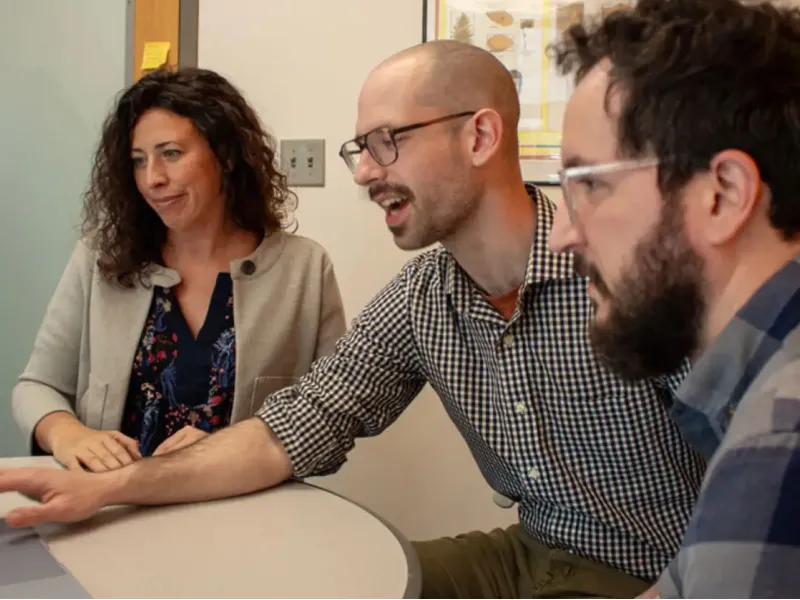 Three people sit around a desk, focused on a laptop. The man in the center gestures enthusiastically, suggesting a collaborative and engaged atmosphere.