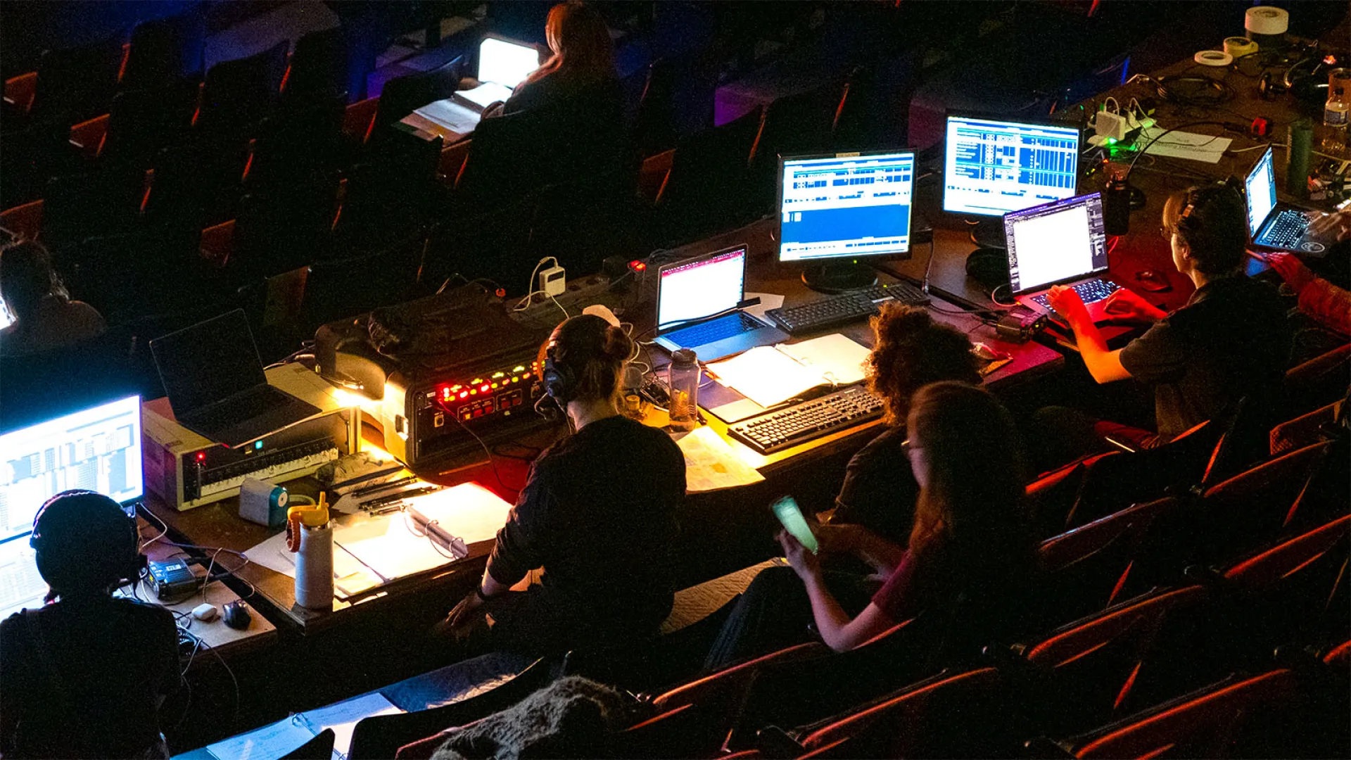 People sitting in a dark theatre, dramatically and colorfully lit by the screens of their monitors and laptops.