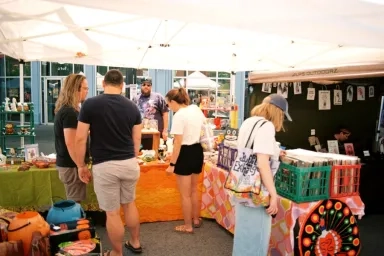 A vibrant outdoor artist market with multiple people browsing and shopping handmade goods and art under a white canopy